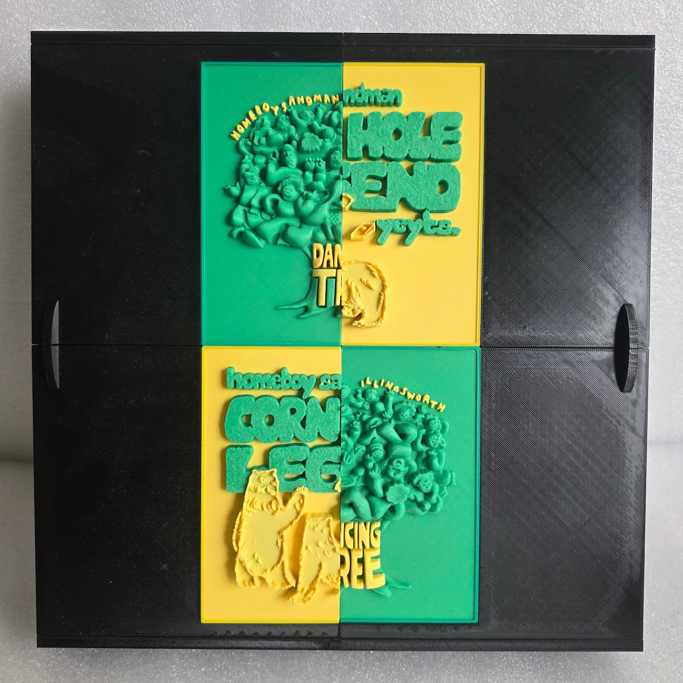 A photograph shows a square, black 3D-printed box or container set against a plain background. The center of the box features a decorative, four-quadrant rectangular plaque with raised, tactile 3D elements printed in vibrant green and yellow filaments. The design is split into four panels with alternating green and yellow backgrounds, forming a cohesive art piece that blends illustrations of a tree packed with stylized cartoon faces and text fragments including "HOMEBOY SANDMAN," "ILLINGSWORTH," "CORN LEG," and "DANCING TREE" across the different sections. The fine, layered texture from the 3D printing process is visible across the entire surface of the black enclosure and the colorful center panels.