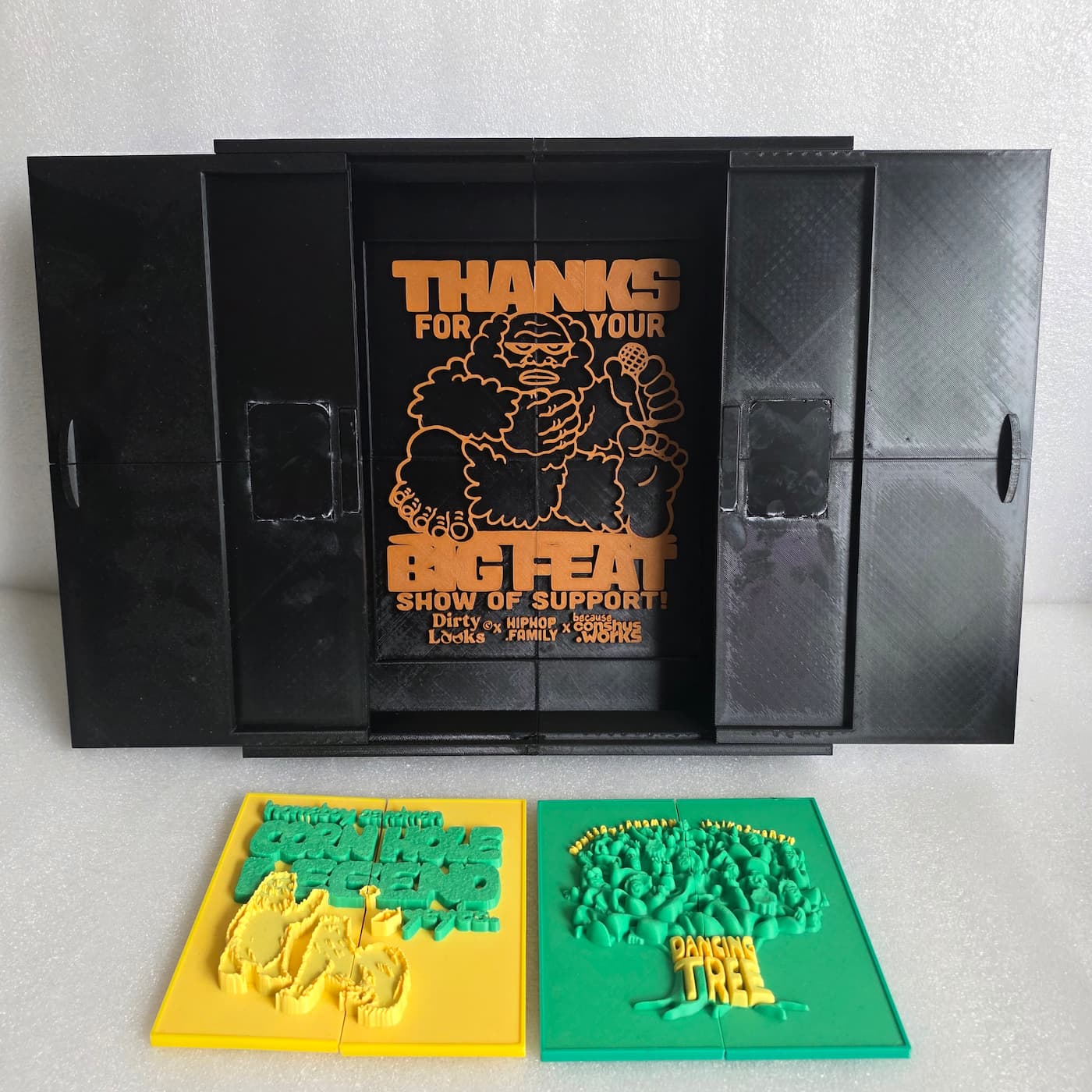 A photograph shows a wide, black 3D-printed box or case with its front doors slid open to reveal an orange and black graphic plate mounted on the inside back wall. The orange graphic features a stylized cartoon depiction of Bigfoot holding a microphone, surrounded by the text "THANKS FOR YOUR BIG FEAT SHOW OF SUPPORT!" along with smaller logos at the bottom for "Dirty Looks," "HIPHOP FAMILY," and "conshus.works". Lying flat in front of the open container are two separate, rectangular 3D-printed plaques featuring raised green and yellow details. The plaque on the left has a yellow background with green text reading "CORN HOLE LEGEND," while the plaque on the right has a green background featuring a tree made of faces and the yellow text "DANCING TREE".