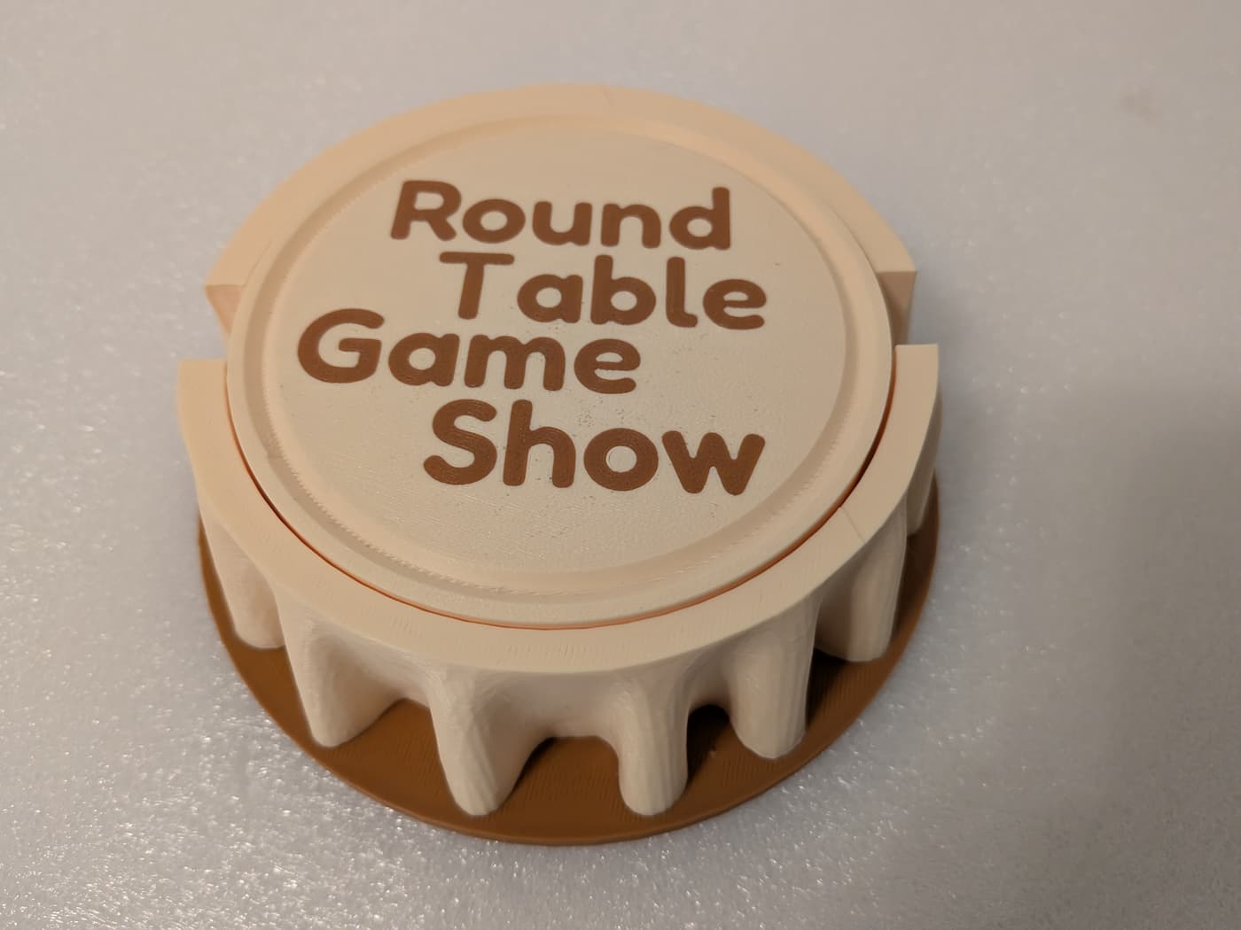 A photograph shows a circular, 3D-printed object styled to resemble a table with a cloth on it. The main body of the cap is printed in a light cream-colored filament, featuring a flat top surface and a fluted, wavy edge that drapes down over a flat, brown circular base. Embossed in the center of the top panel is the text "Round Table Game Show" stacked in four rows, rendered in a matching brown, rounded sans-serif font. The distinct, fine layer lines characteristic of 3D printing are subtly visible across the entire textured plastic surface.