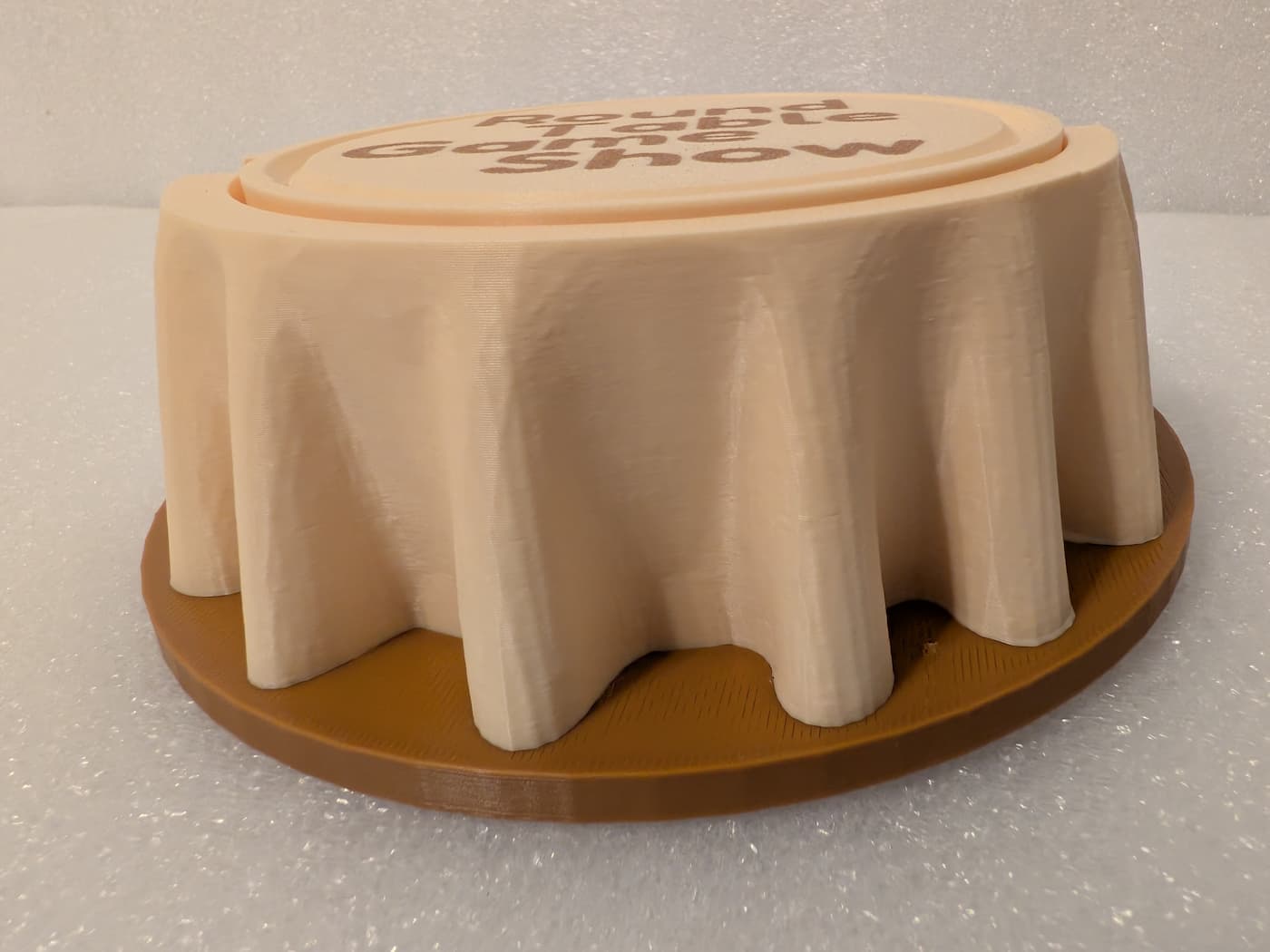 A photograph captures a low-angle, side view of a 3D-printed plastic object resembling a round table draped with a tablecloth. The main structure is printed in a light cream-colored filament, showing deep, sculpted folds that drape down toward a flat, circular brown plastic base. The slightly angled top surface reveals a portion of brown text that reads "Round Table Game Show". The entire surface displays the fine, horizontal layer lines from the 3D printing process, and the object is set against a textured, light-colored background.