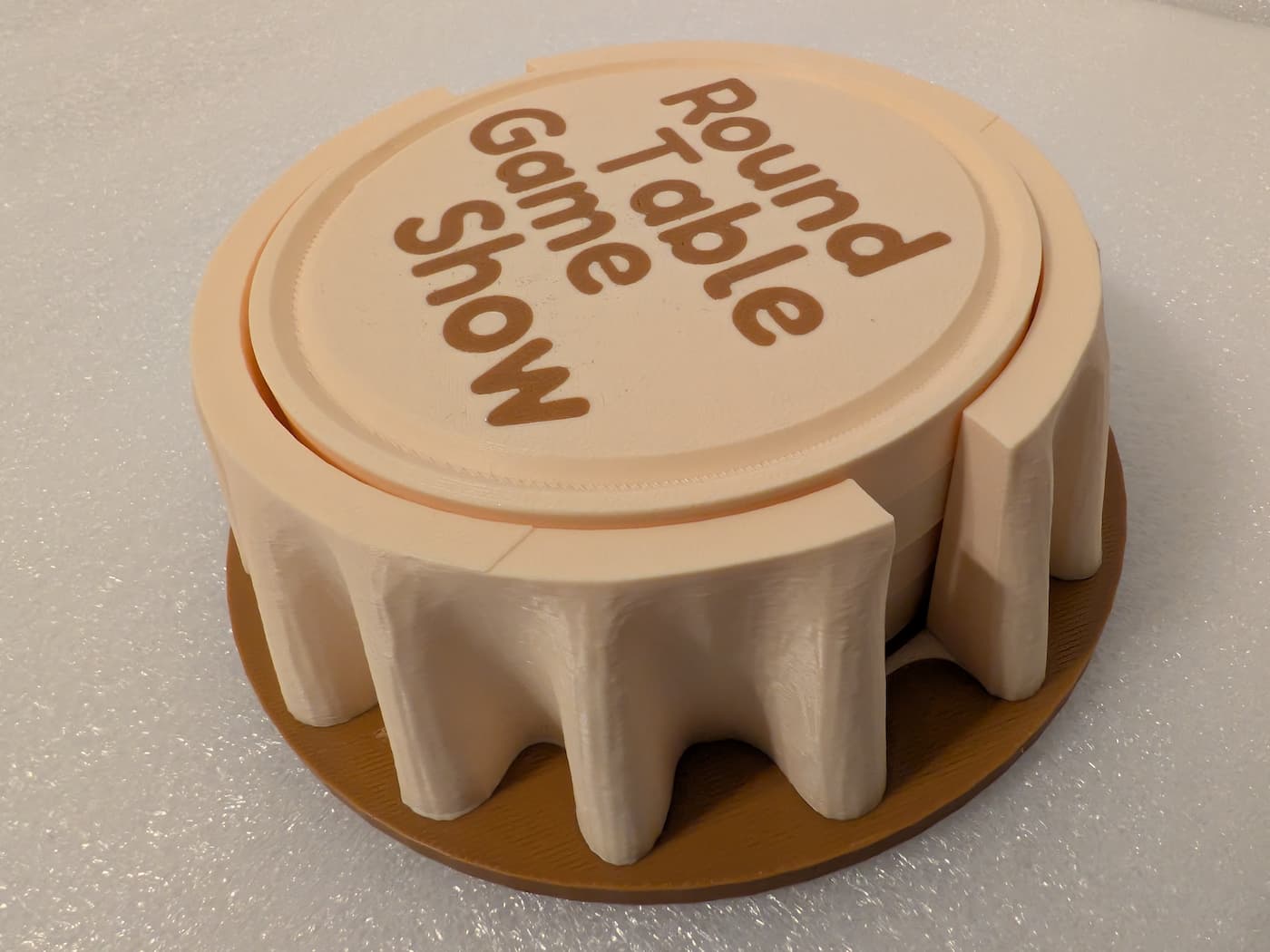 A photograph captures a high-angle, side view of a circular, 3D-printed set of coasters and a holder designed to resemble a round table with a hanging tablecloth. The upper structure is printed in a light cream-colored filament, featuring deep drapery folds along its outer edge that descend onto a flat, circular brown base. The top surface features a raised, recessed circular insert with the text "Round Table Game Show" printed in matching brown, rounded letters across four lines. Fine, horizontal layer lines from the 3D printing process are visible throughout the entire textured plastic piece, which is resting on a smooth, off-white surface.
