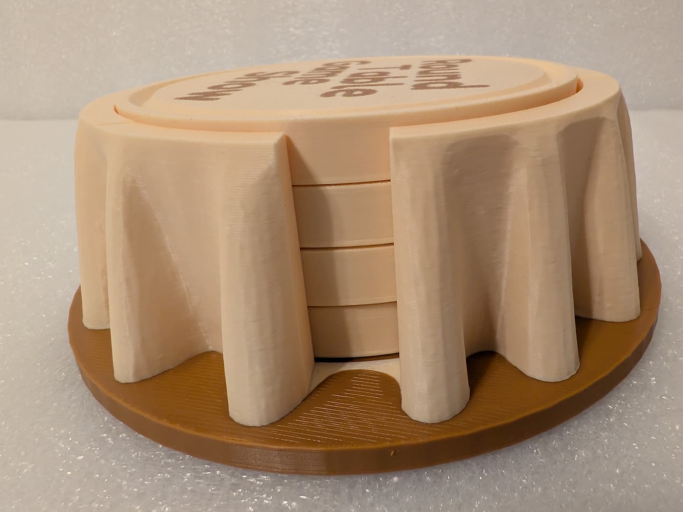 A photograph captures a low-angle, side view of a 3D-printed coaster holder and coaster set designed to look like a round table. The main holder is printed in a light cream-colored filament, featuring deep, sculpted folds that resemble a hanging tablecloth draped over a flat, circular brown plastic base. A large vertical opening in the side of the holder reveals a stack of four matching cream-colored circular coasters stored neatly inside. The top surface of the top coaster shows a portion of text that reads "Round Table Game Show". The entire surface displays the fine, horizontal layer lines from the 3D printing process against a textured, light background.