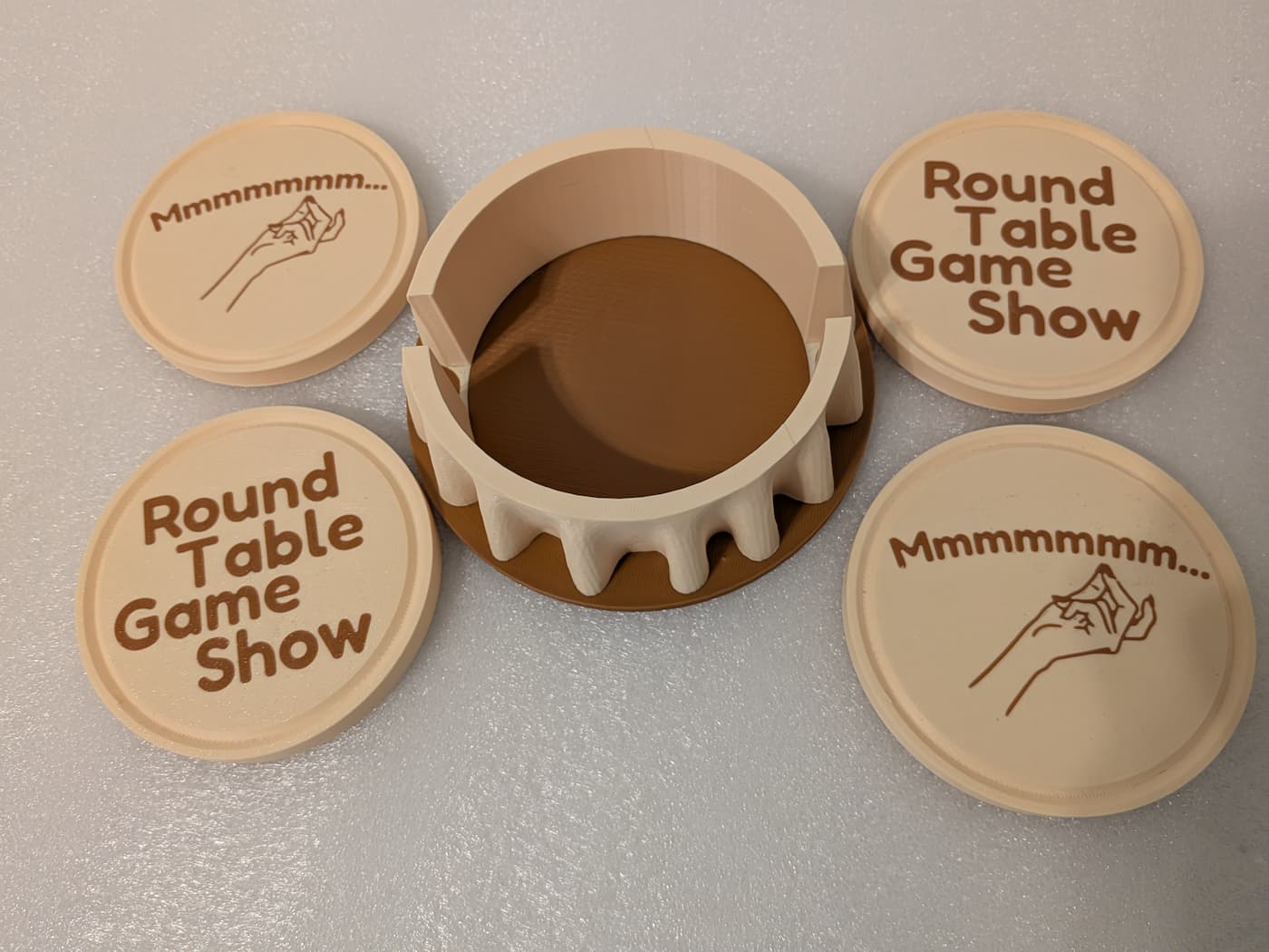 A high-angle photograph shows an empty, circular 3D-printed coaster holder surrounded by four matching drink coasters arranged on a smooth, off-white surface. The holder features a light cream-colored upper rim with decorative, wavy folds resembling a draped tablecloth that sits over a flat, brown circular base. The four cream-colored circular coasters are arranged in pairs. Two of the coasters feature the text "Round Table Game Show" printed in a rounded brown font across four lines. The other two coasters depict a simple line drawing of a hand performing a chefs kiss gesture accompanied by the brown text "Mmmmmmm...". Fine, horizontal layer lines from the 3D printing process are visible across all pieces.