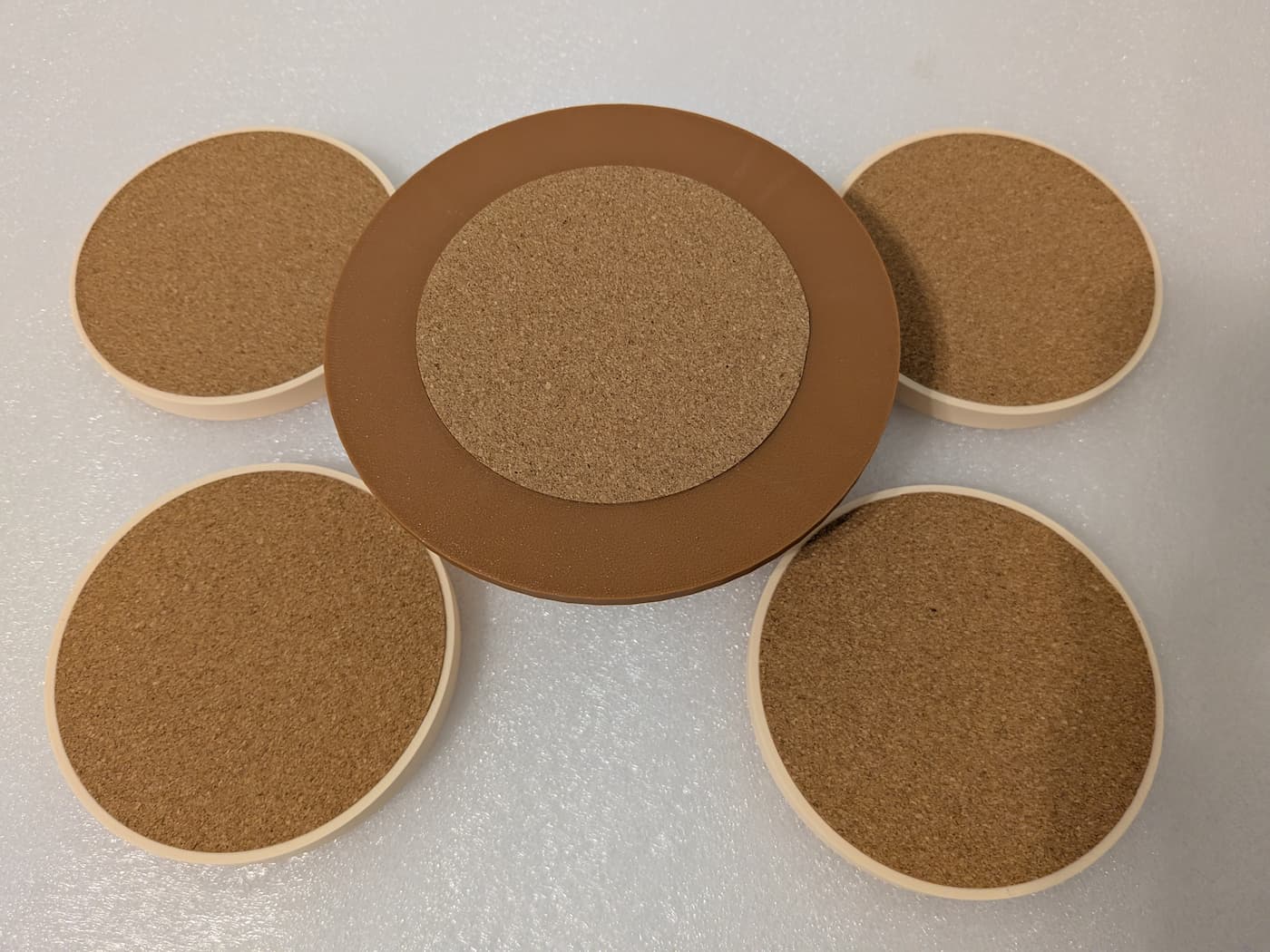 A high-angle photograph shows the undersides of a circular 3D-printed coaster holder and four matching drink coasters arranged face-down on a smooth, off-white surface. In the center, the large, flat brown bottom of the holder features a circular, textured brown cork pad adhered to its middle. Surrounding the holder, the four coasters are placed face-down, each revealing a thick, circular cork backing insert fitted flush within its thin, cream-colored plastic rim. The fine, circular print lines are visible on the exposed plastic portions, highlighting the 3D-printed construction of the set.