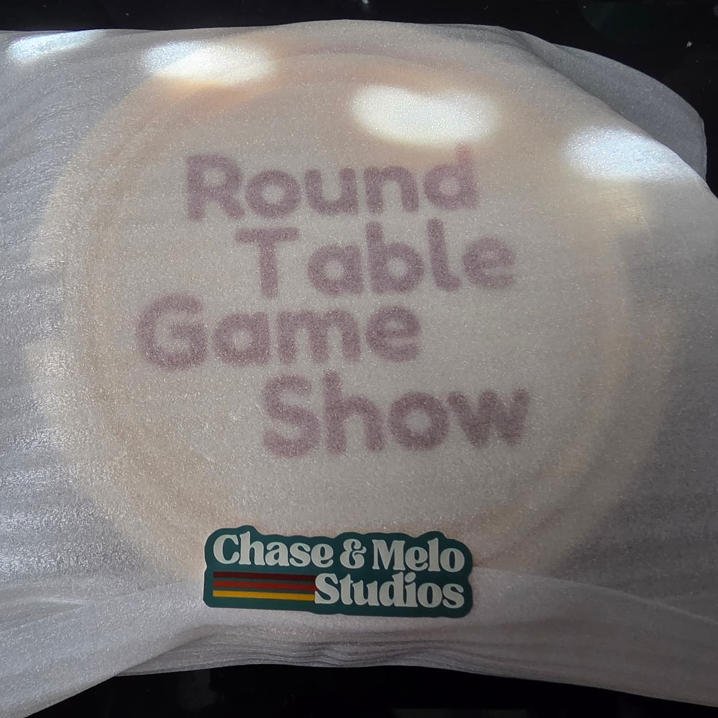 A close-up photograph shows a circular item wrapped inside a translucent, textured white protective foam sheet. Visible through the thin foam wrapper is the brown, stacked text "Round Table Game Show" printed on the object inside. Affixed to the outside of the protective packaging near the bottom is a small sticker displaying the "Chase & Melo Studios" logo, featuring dark green, retro-styled text accompanied by three horizontal accent stripes in dark red, orange, and yellow.