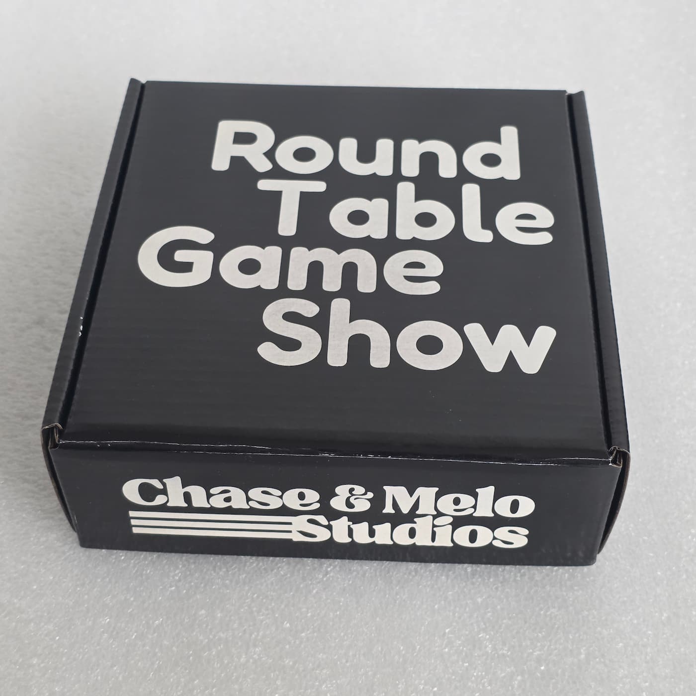 A high-angle photograph shows a square, black cardboard mailer box resting on a textured, light grey surface. Printed in a bold, rounded white or silver font across four lines on the top lid of the box is the text "Round Table Game Show". The front side panel of the box features the white "Chase & Melo Studios" logo, which consists of the brand name in a stylized, retro-inspired serif font accompanied by three horizontal accent lines on the left.