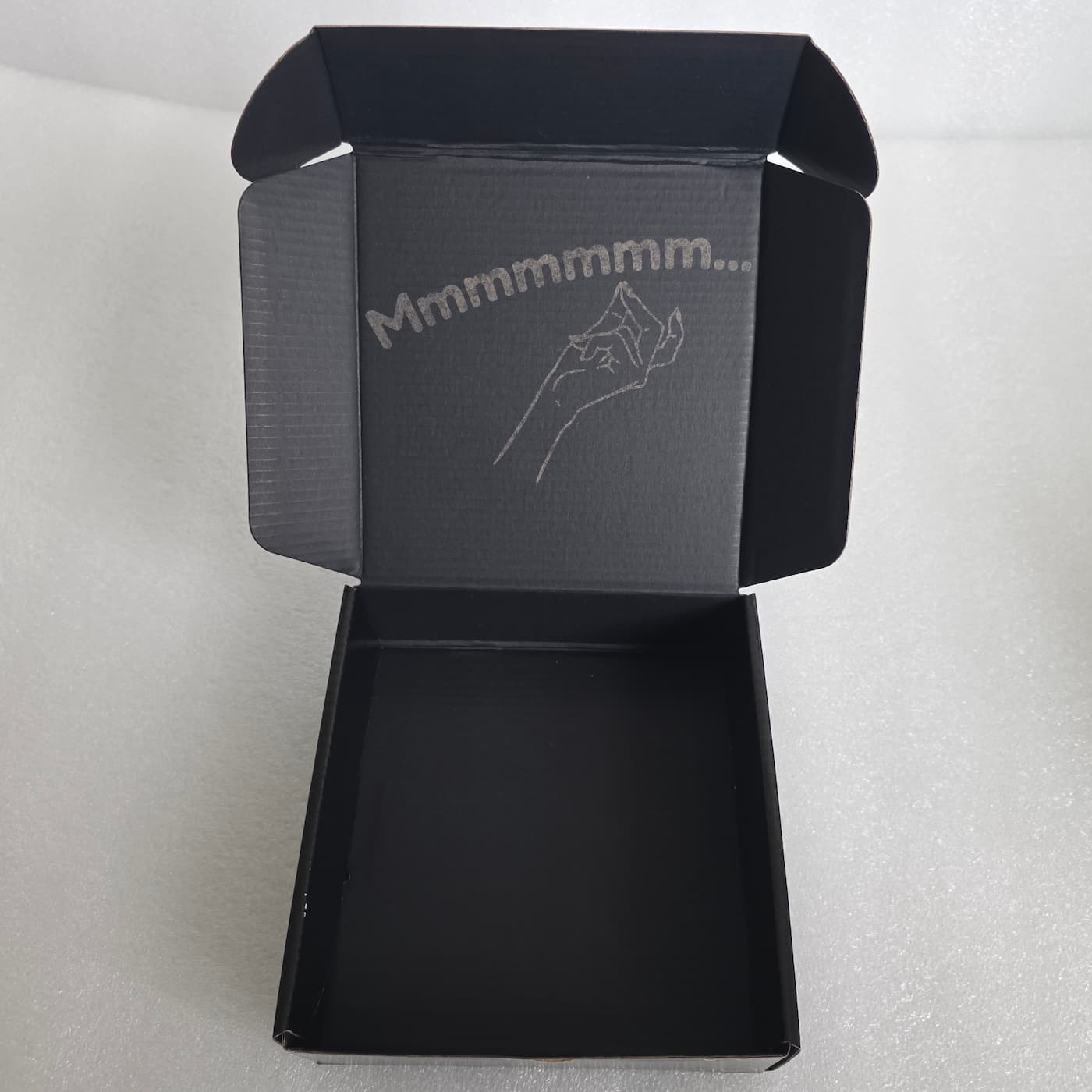 A high-angle photograph shows an empty, open black cardboard mailer box resting on a textured, light grey surface. On the inside of the top lid, a grey line drawing depicts a hand performing a chefs kiss gesture, arched beneath the printed text "Mmmmmmm...". The deep main compartment of the box stands open and empty in the foreground.