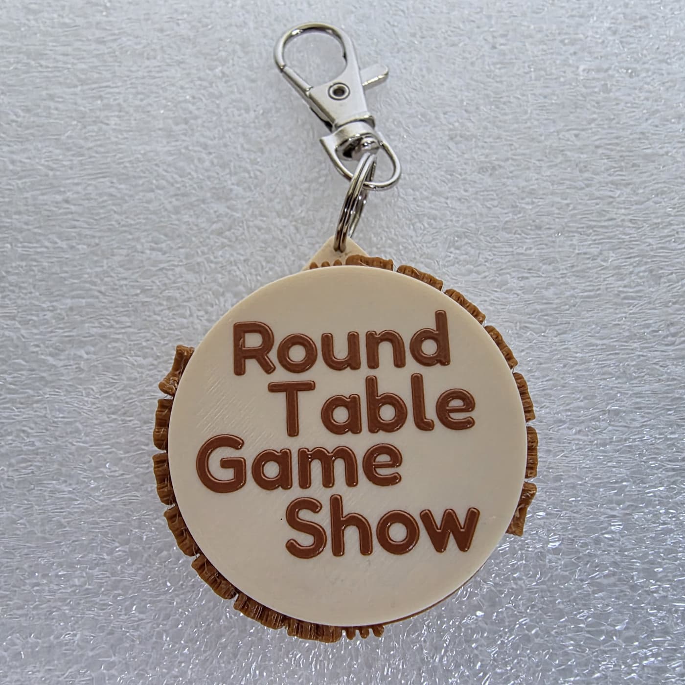 A photograph captures a circular, 3D-printed keychain resting on a textured, white protective foam surface. The main body of the keychain features a smooth, cream-colored circular face embossed with the text "Round Table Game Show" stacked across four lines in a rounded, brown font. Layered behind the cream disc is a slightly larger, brown 3D-printed base with a rough, textured ridge extending past the border. A small loop at the top connects to a silver metal split ring and a lobster claw clasp.