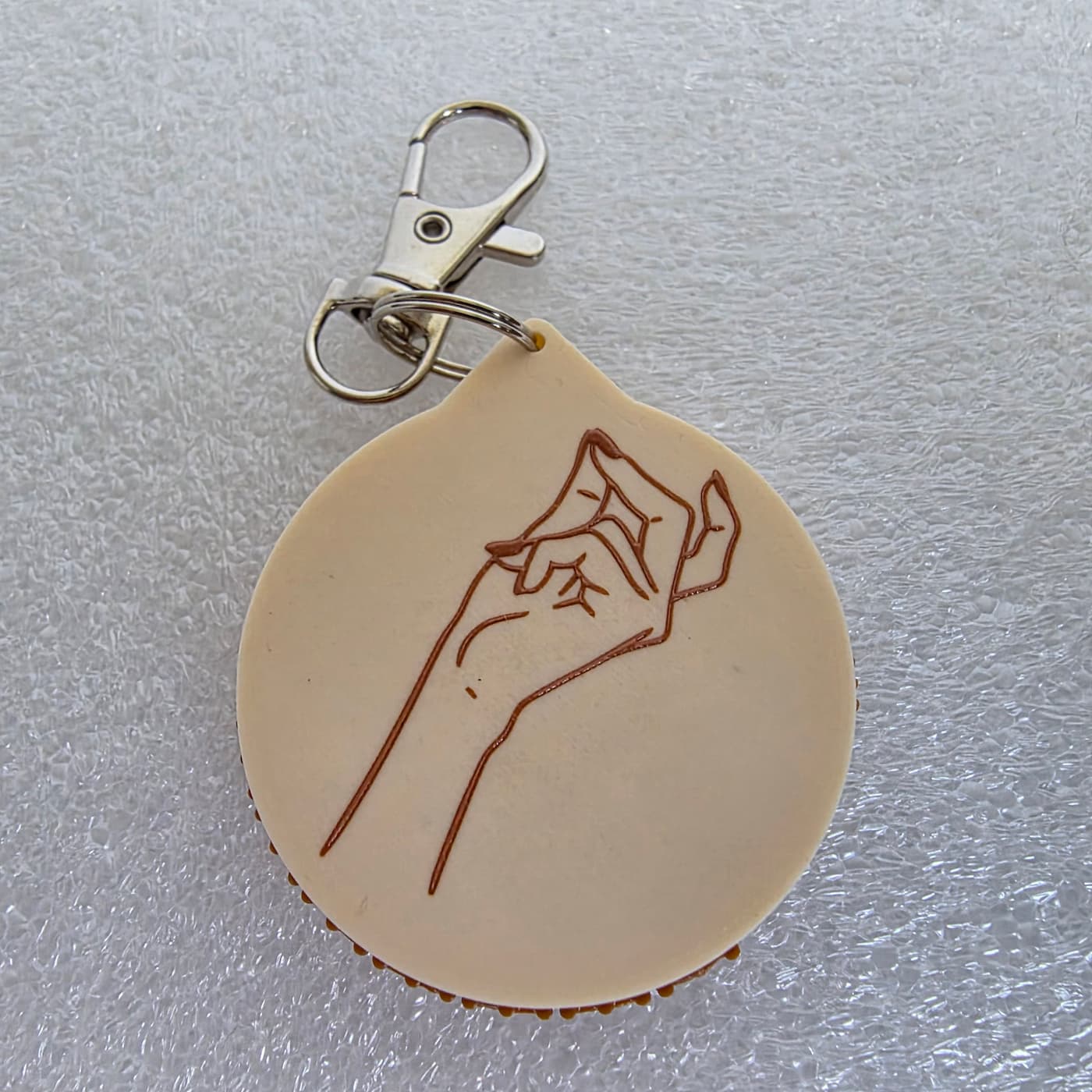 A photograph captures the reverse side of a circular, 3D-printed keychain resting on a textured, white protective foam surface. The smooth, cream-colored plastic face features a simple brown line drawing of a hand performing a chefs kiss gesture. A slightly larger brown plastic backing layer with small textured ridges is faintly visible along the bottom edge. At the top, a built-in loop connects to a silver metal split ring and a lobster claw clasp attachment.