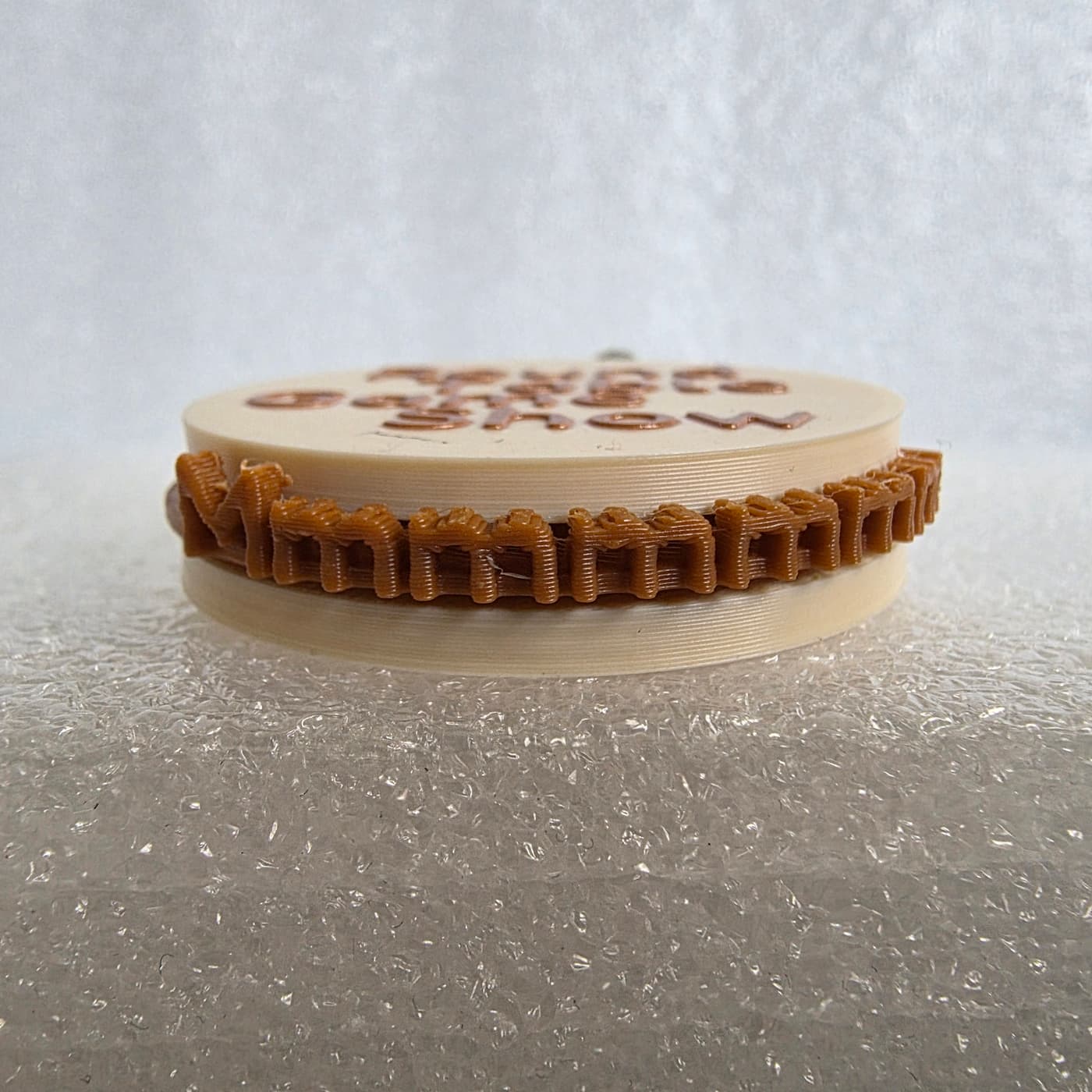 A photograph captures a low-angle, side view of a circular, sandwich-style 3D-printed keychain resting on a textured, white protective foam surface. The top and bottom layers are made of smooth, cream-colored plastic discs, with out-of-focus brown text partially visible on the upper surface. Sandwiched tightly between the two cream layers is a distinct middle band made of brown filament, intricately 3D-printed to form a repeating pattern of the letters "m" spelling out "Mmmmmmm..." around the entire circumference. The fine, horizontal layer lines from the manufacturing process are clearly visible across all edges of the plastic piece.