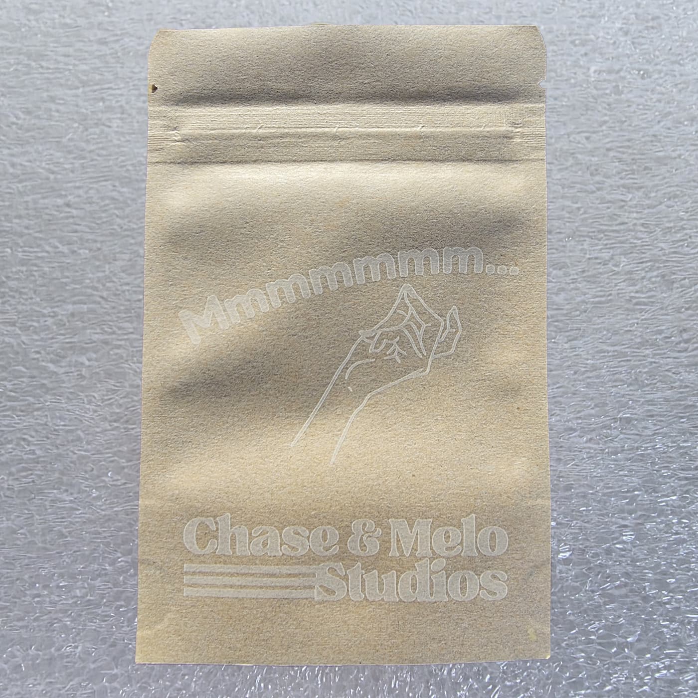 A front-facing photograph shows a small, rectangular brown kraft paper pouch with a resealable zipper top, resting on a textured, white protective foam surface. Printed in a subtle, translucent white ink on the center of the bag is a line drawing of a hand performing a chefs kiss gesture, beneath the arched text "Mmmmmmm...". The bottom of the pouch features the white, retro-style "Chase & Melo Studios" logo, including its three distinct horizontal accent stripes on the left.